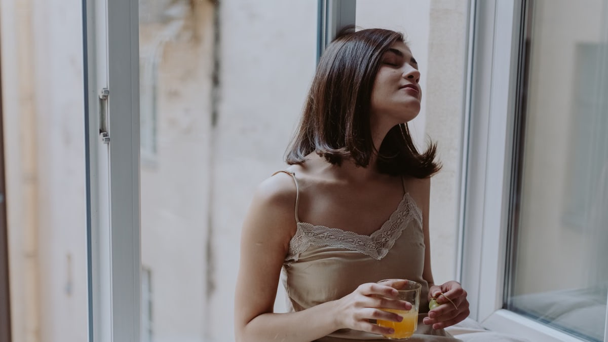Woman in a spaghetti strap dress savoring orange juice on a window sill, indoors.